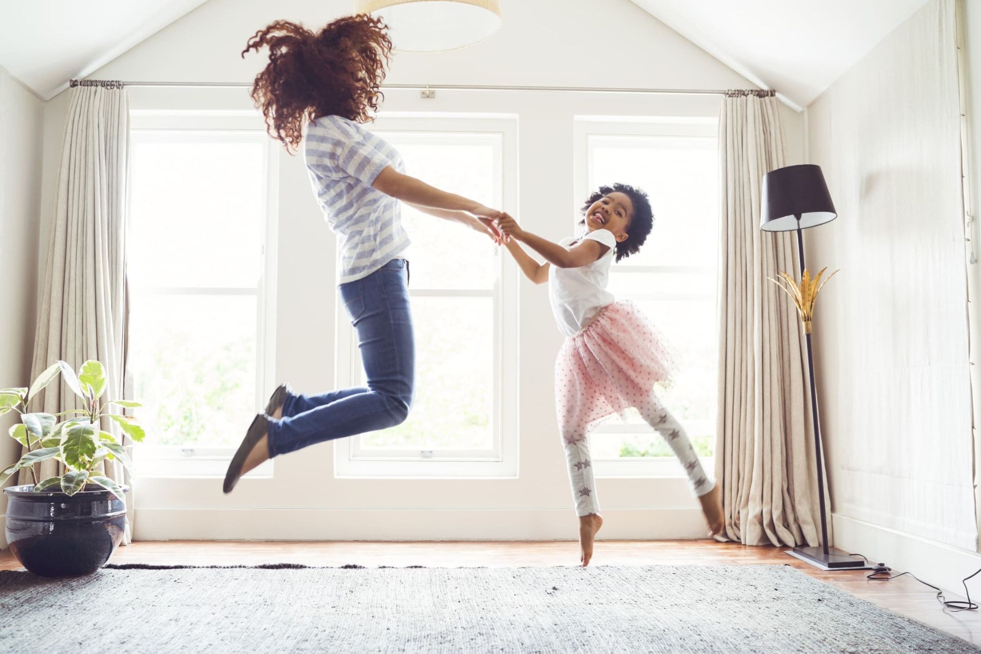 A caregiver and a young girl jumping and holding hands in a sunlit living room